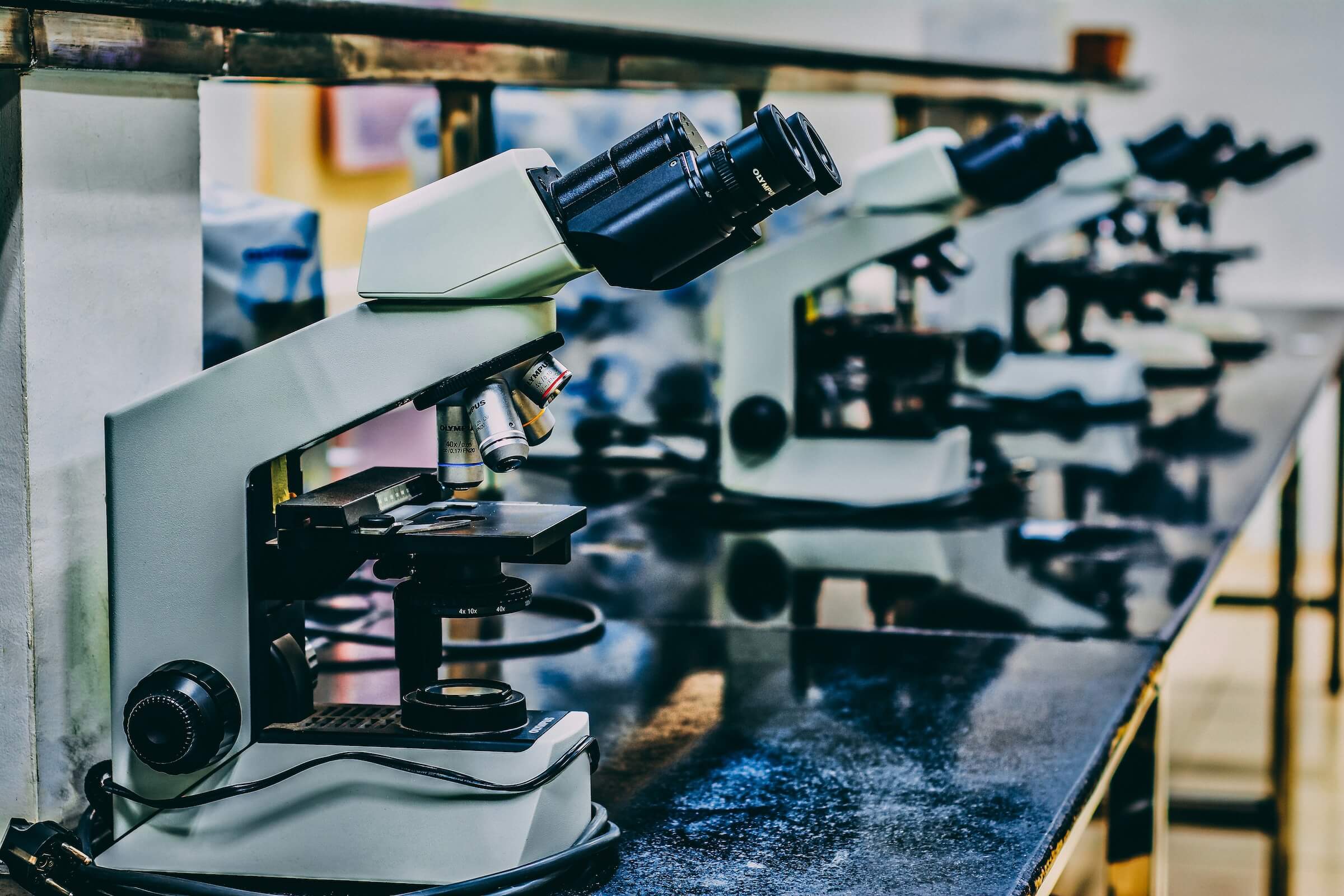 Rząd białych mikroskopów ustawionych na czarnym blacie w laboratorium. Pierwszy mikroskop jest ostry, a kolejne stopniowo rozmywają się w oddali